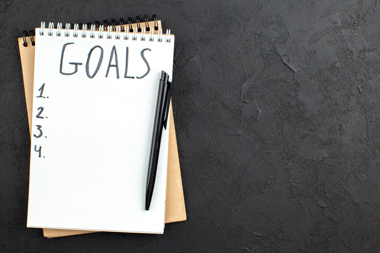 A notepad labeled "GOALS" with a list numbered from 1 to 4, and a black pen placed on top, lying on a dark textured surface.